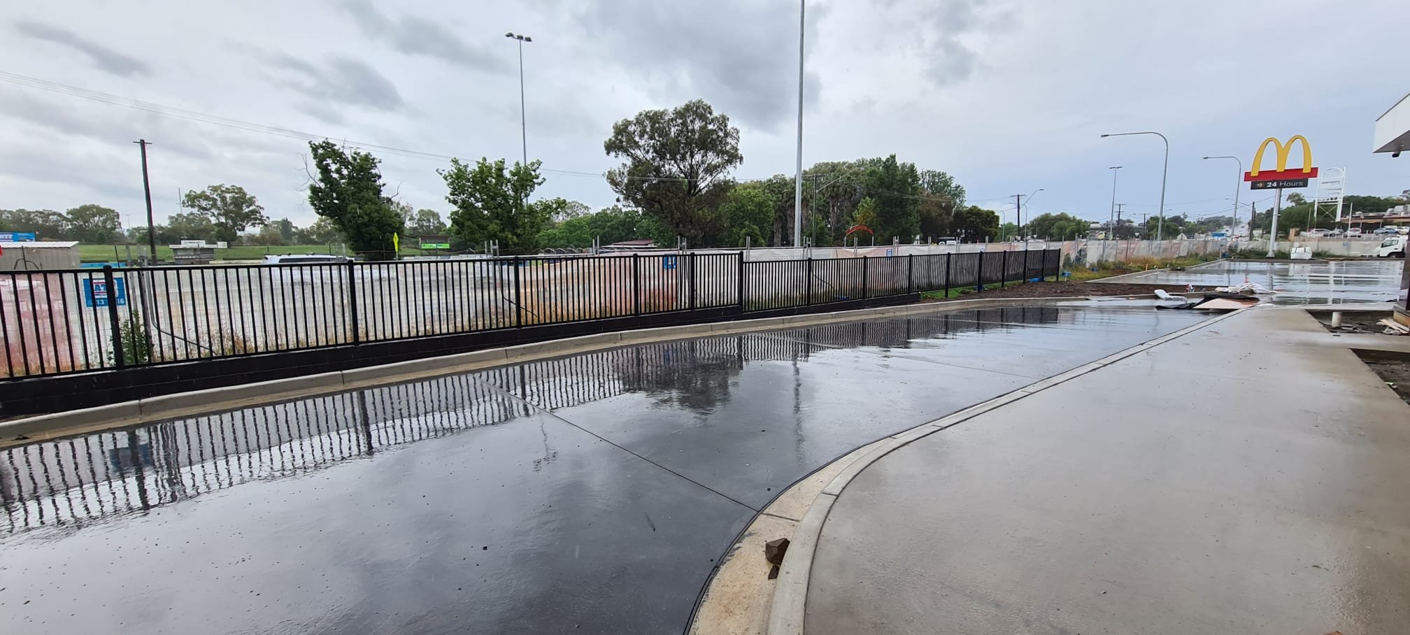 A wet sidewalk outside a McDonald's restaurant after rain, with puddles reflecting the cloudy sky, and a black metal fence separating the sidewalk from a grassy area behind it. The McDonald's sign is visible in the background.