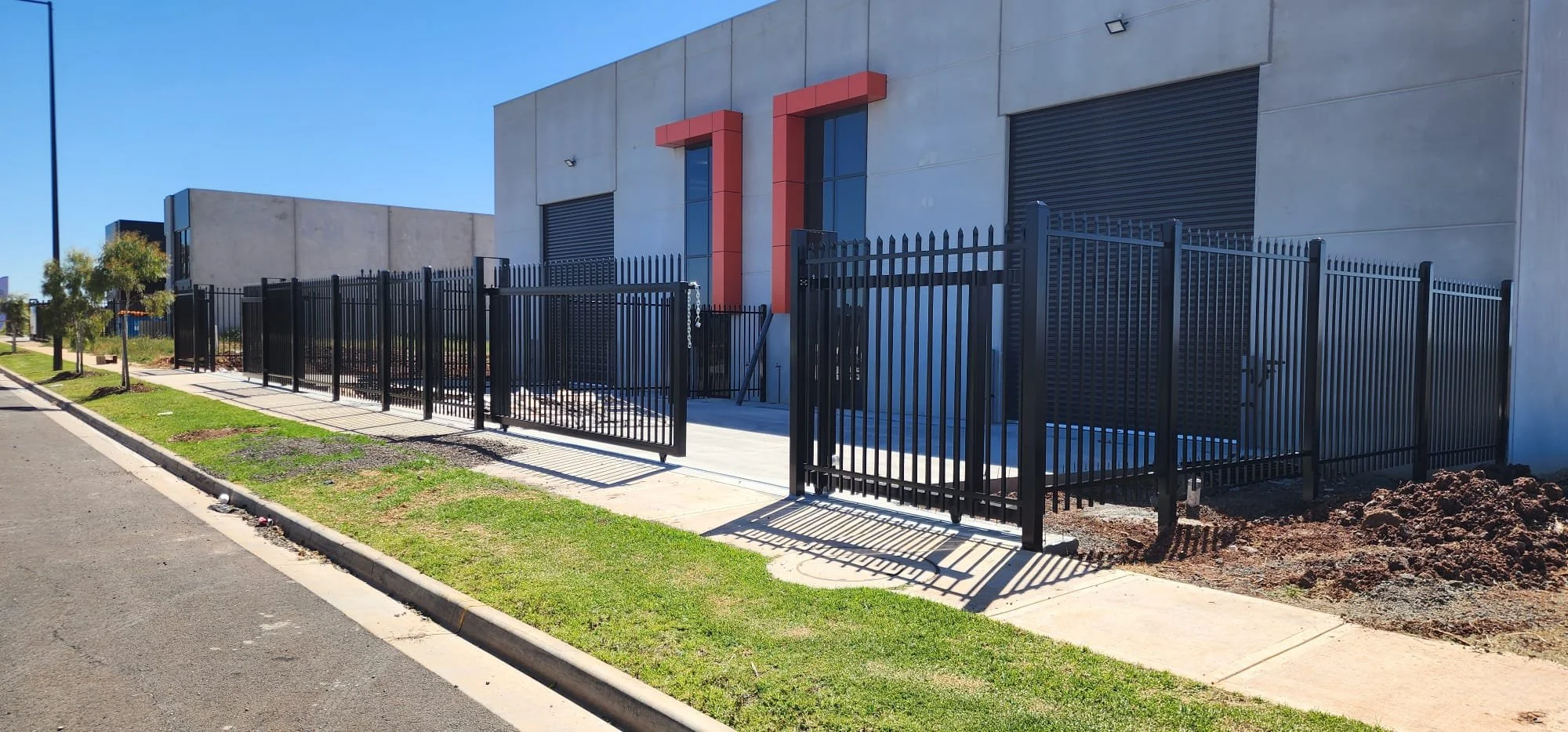 Modern building with black metal fence and gate, concrete sidewalk, green grass, and small trees along the street under a clear blue sky.