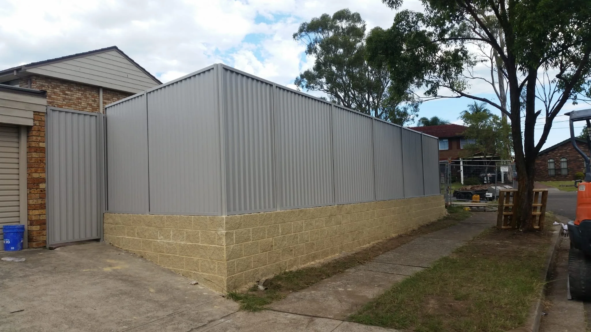 A construction site showing a newly erected metal fence with a brick foundation surrounding a property, with a sidewalk, trees, and nearby houses in the background.