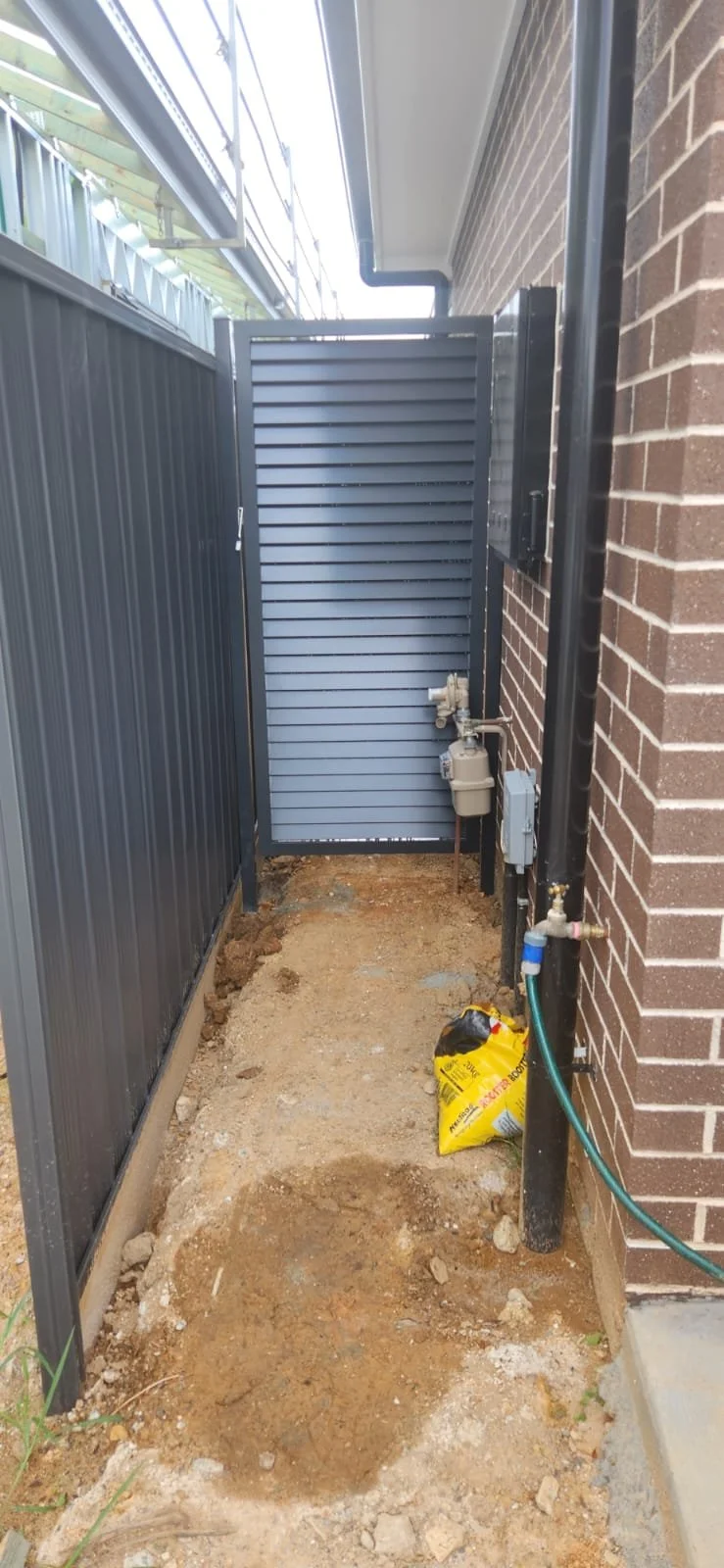 Side yard with dirt ground, a brick wall, and a metal utility box connected to piping. A garden hose is on the ground next to the brick wall, and a yellow bag of soil is present. A metal gate is at the end of the space.