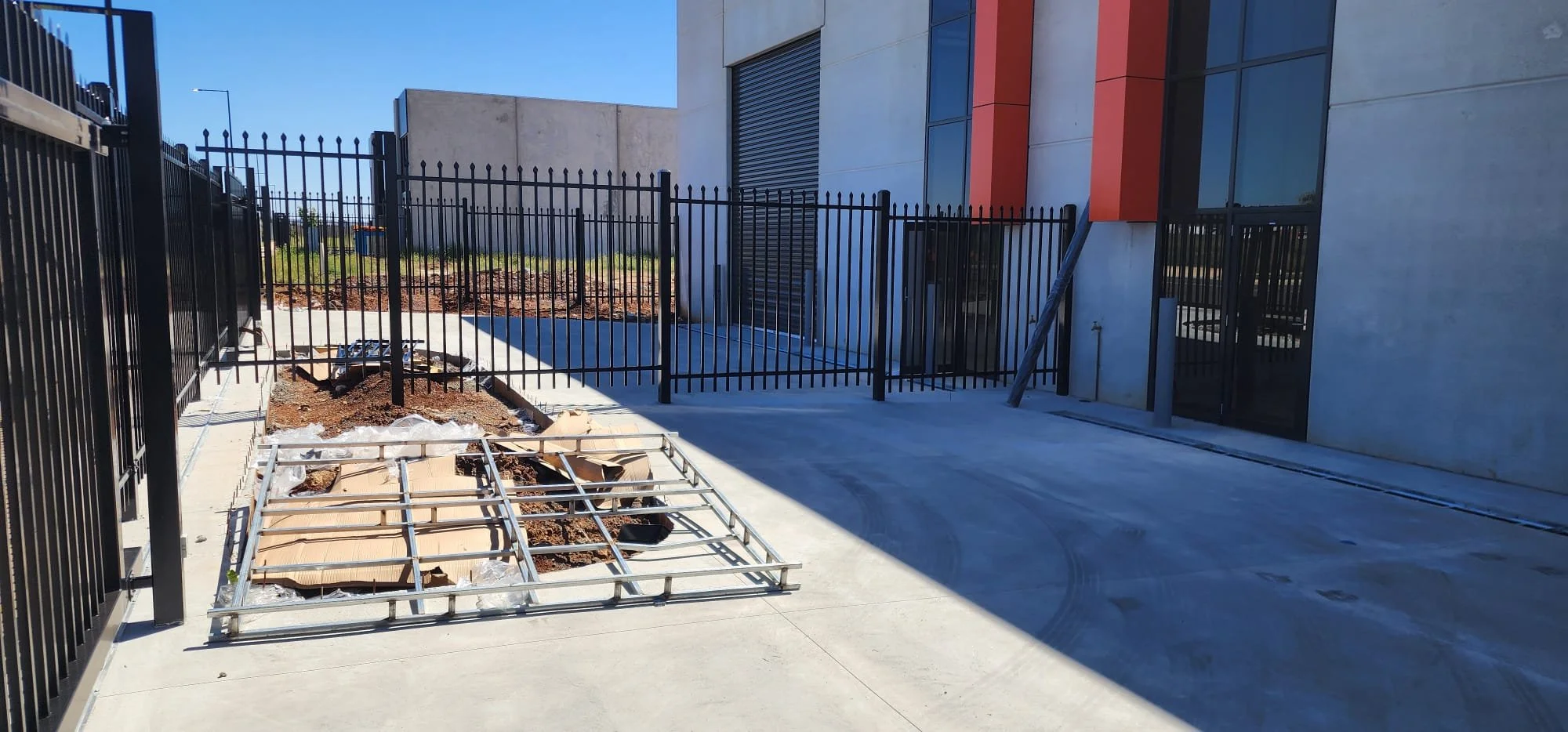 Construction site with black metal fence, concrete pavement, and building with large windows and garage door, during daytime.