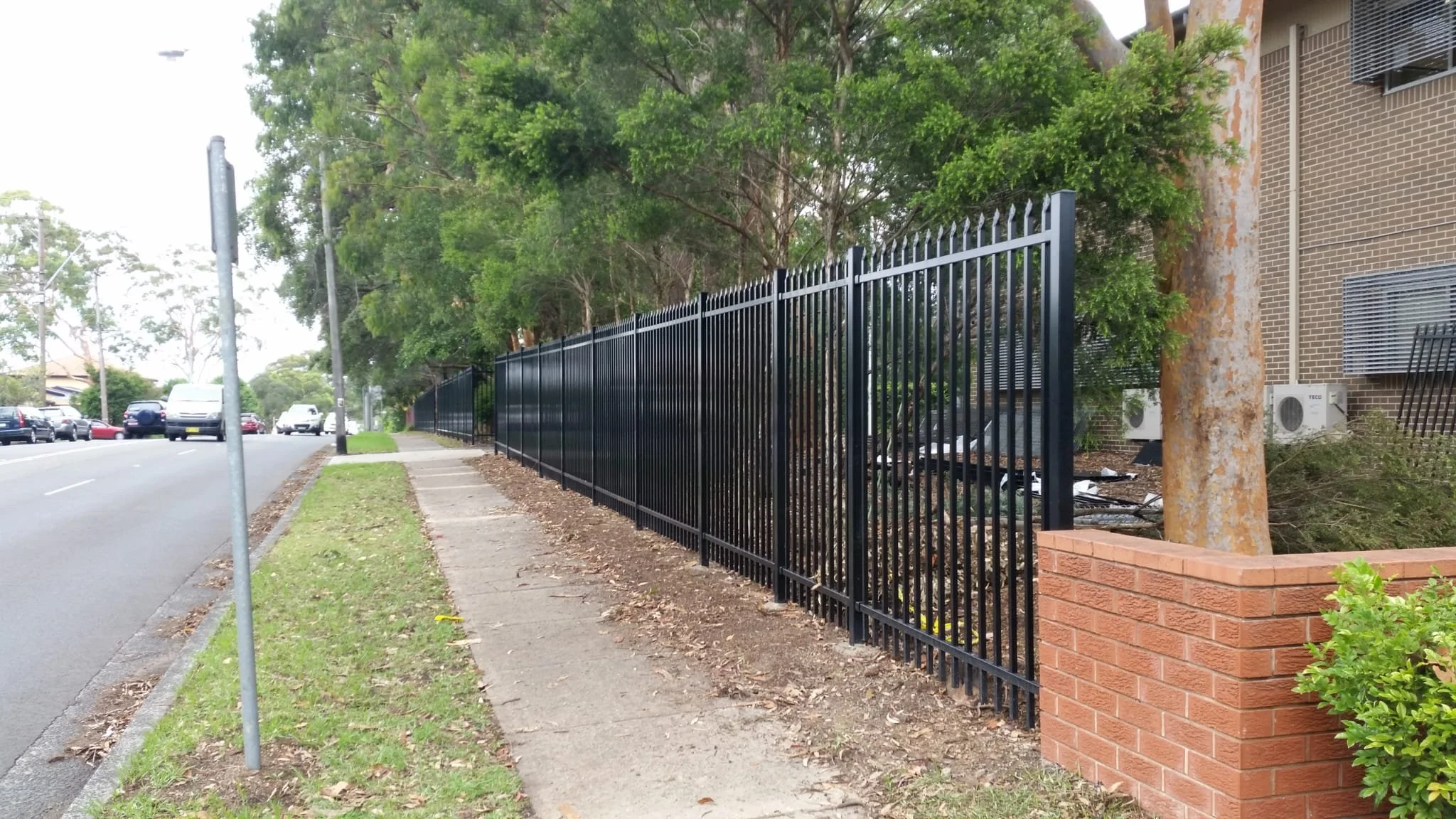 A sidewalk runs along a street with parked cars on the left side. To the right, there is a tall black metal fence with vertical bars, running parallel to the sidewalk. Behind the fence, there are trees with green foliage and some brick and concrete b