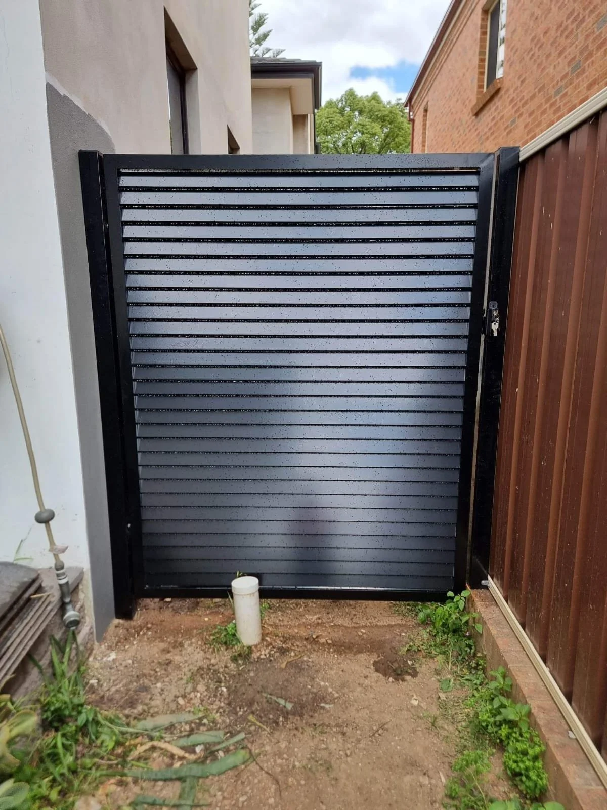 Black metal gate with horizontal slats, in a narrow backyard space between a concrete house and a wooden fence, with green plants and dirt ground.
