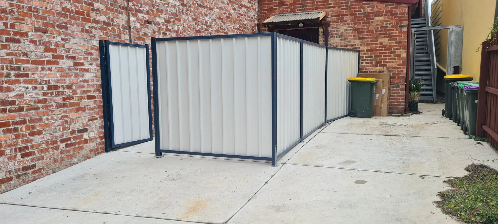 A small outdoor enclosed trash area with white metal fencing, located next to a brick wall and concrete ground, with trash bins and a staircase in the background.