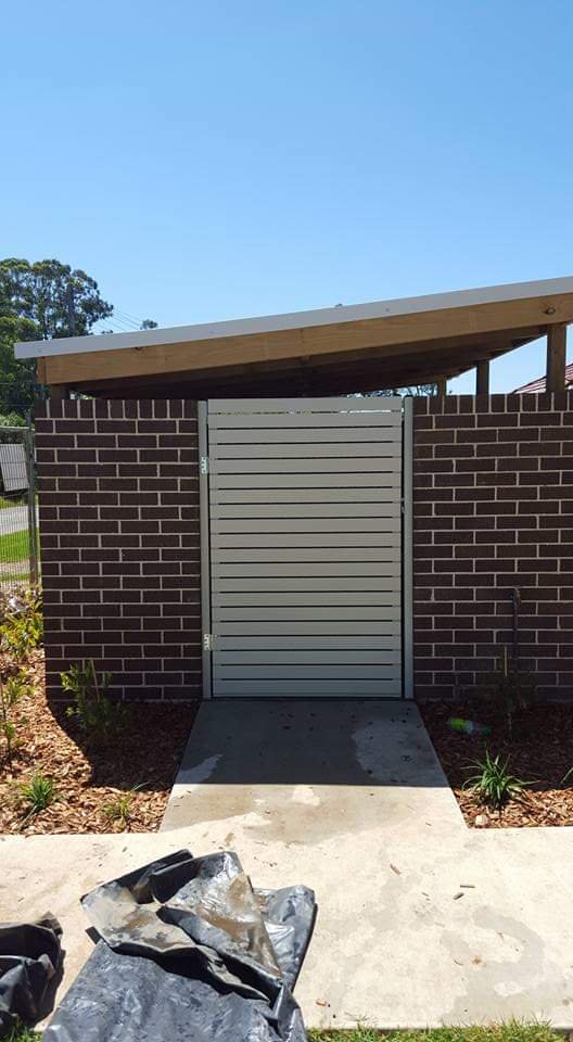 A small backyard gate made of gray horizontal slats, set within a brick wall with a concrete pathway leading up to it, surrounded by small plants, under a clear blue sky.