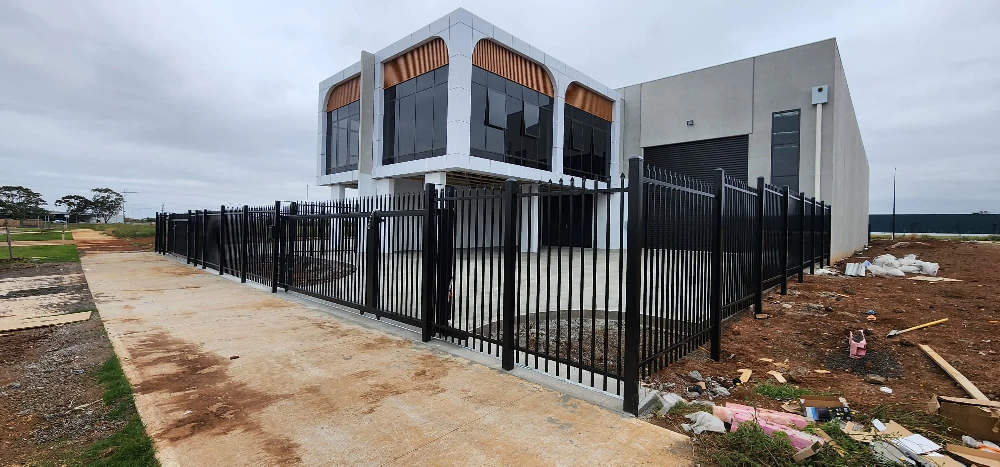 Modern two-story house with large glass windows, surrounded by a black metal fence, under cloudy sky, with construction debris on the ground.