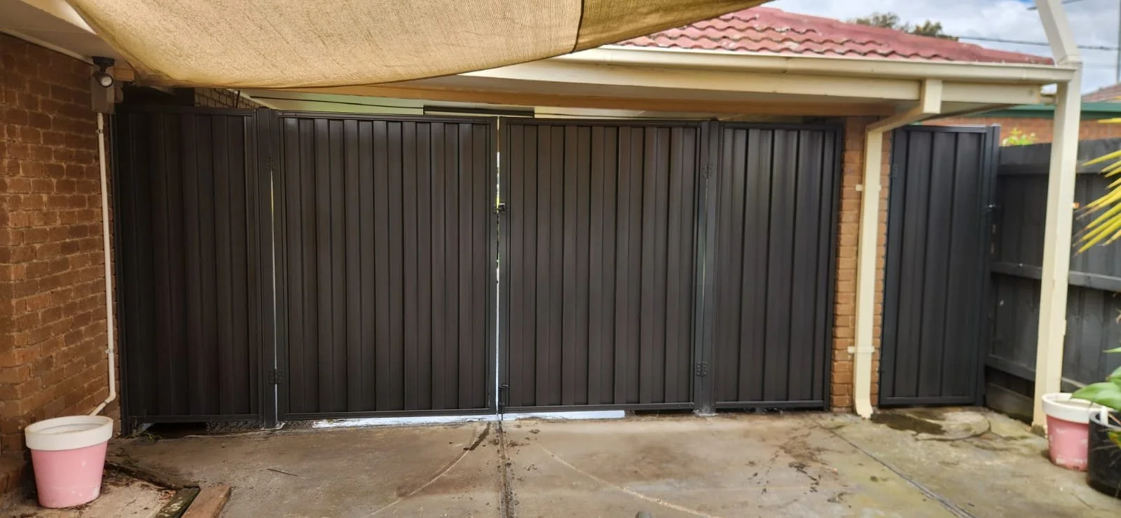 A closed black metal gate on a concrete driveway, with a brick wall on the left and a wooden fence on the right, part of a residential carport area.