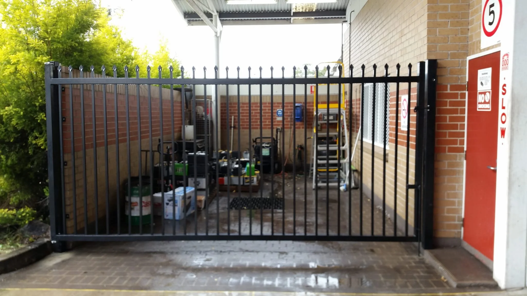 Locked black metal gate blocking a small outdoor utility area with tools and storage containers, next to a brick building with a fire escape and no smoking sign.