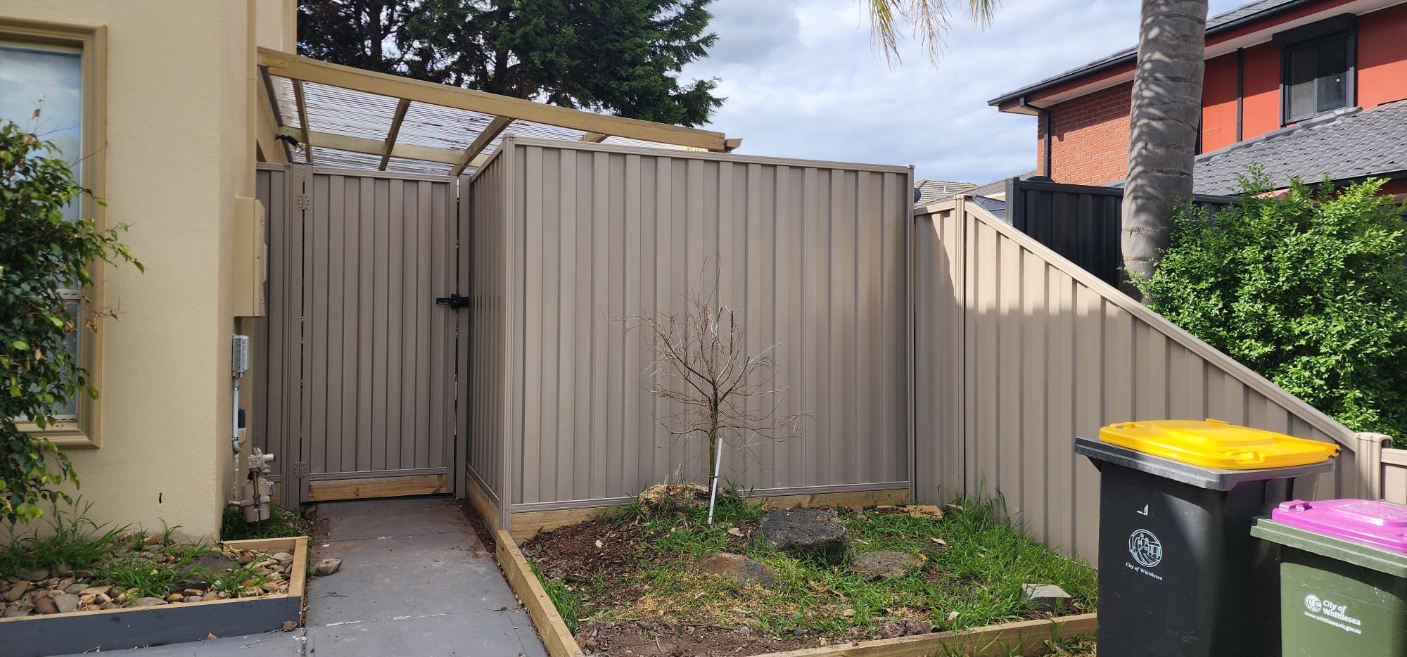 Backyard with beige metal fence, small leafless tree, two trash cans with yellow and pink lids, and green bushes, with neighboring house and palm tree in the background.