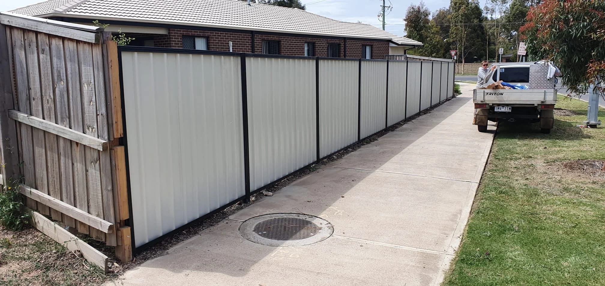A sidewalk next to a new white fence with black frames, with a man standing in the back near a pickup truck loaded with household items, and a brick building in the background.