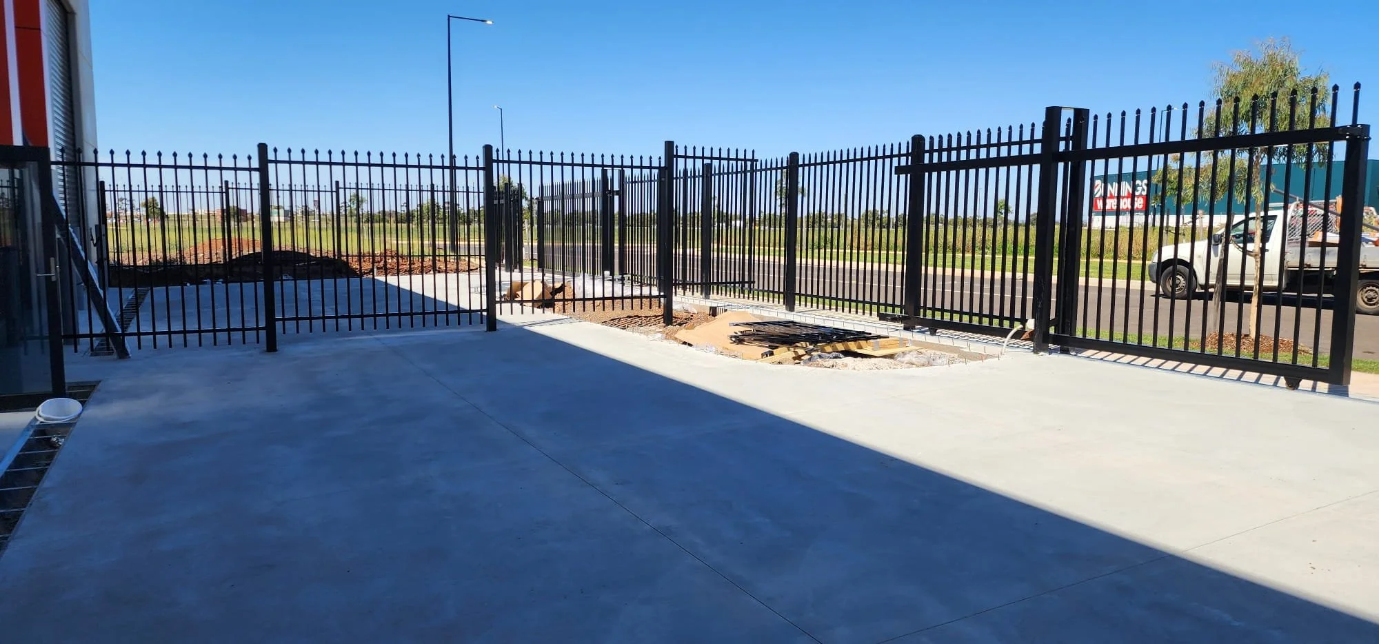 Black metal fence with gates, construction materials and dirt behind it, concrete floor in the foreground, blue sky, parked cars and signage in the background.