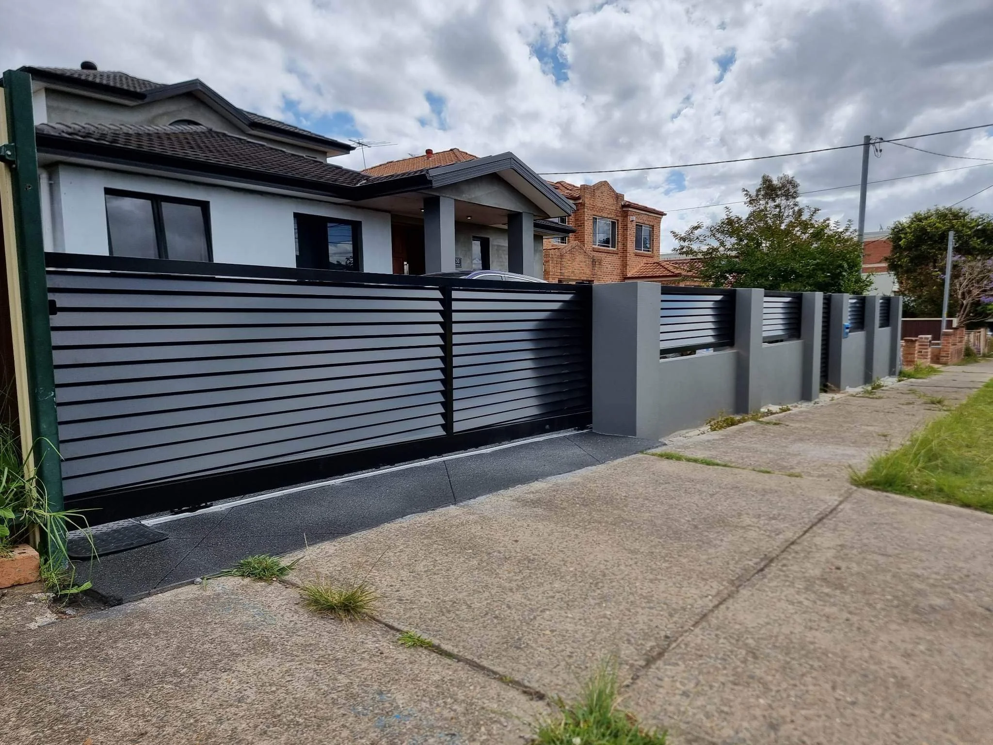 Modern house with a metal fence and gate, sidewalk, cloudy sky, and neighboring houses.