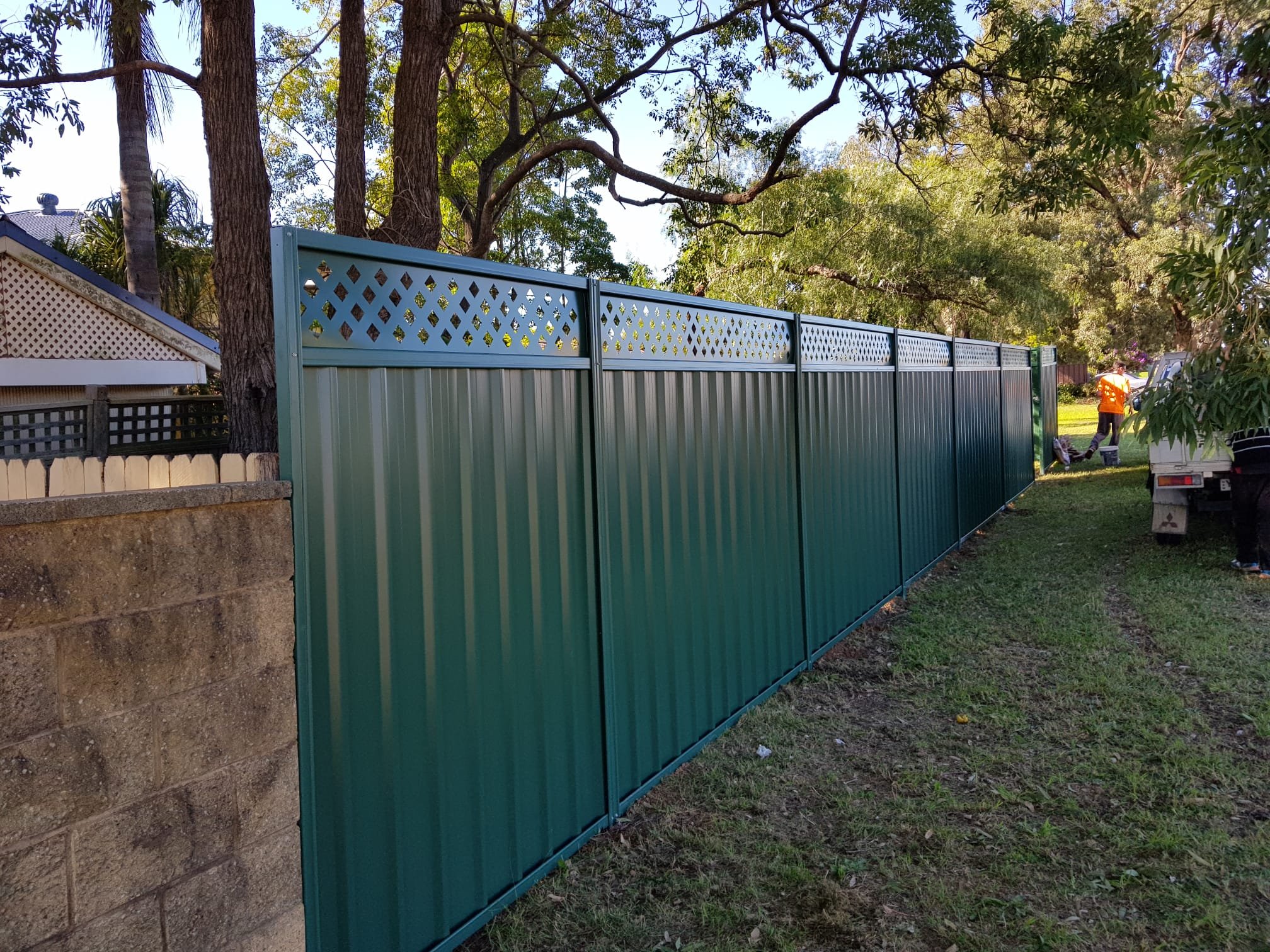 New green metal fence installation on a residential property with workers in the background, trees, and a vehicle nearby.