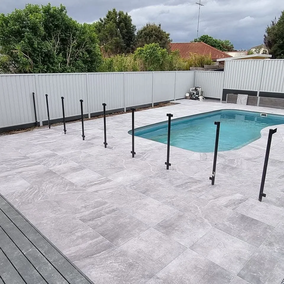 A backyard with a rectangular swimming pool surrounded by grey tiles and a white fence. The pool has a barrier around it with black posts, and there is some equipment in the corner of the yard. The sky is cloudy with dark clouds.