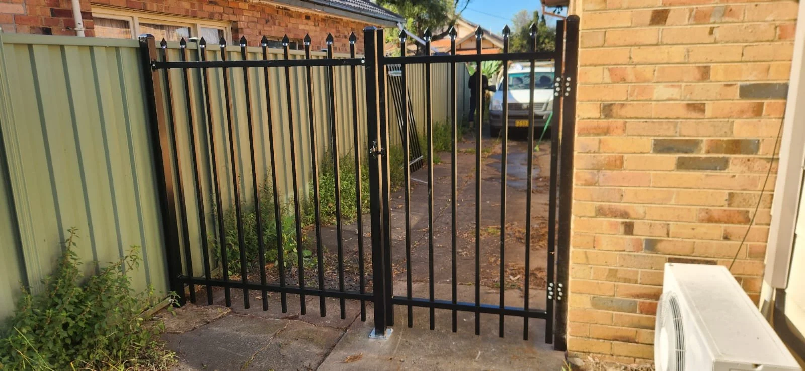 A black metal gate in front of a backyard with a concrete path, some greenery, a parked white van, and a brick house wall with an air conditioning unit.