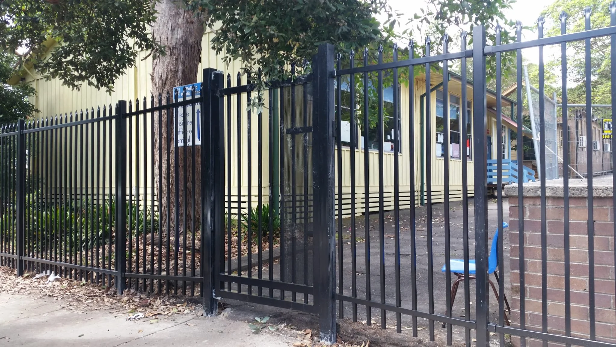 View of a black metal fence surrounding a small playground or park area with a yellow building, trees, and a blue chair visible inside the enclosed space.