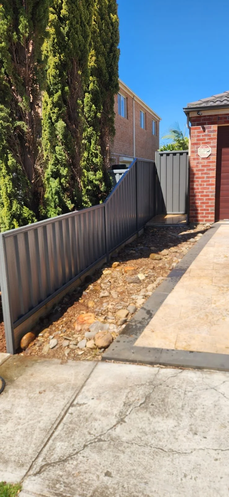 A backyard corner with a metal fence, brick house wall, and tall evergreen trees.