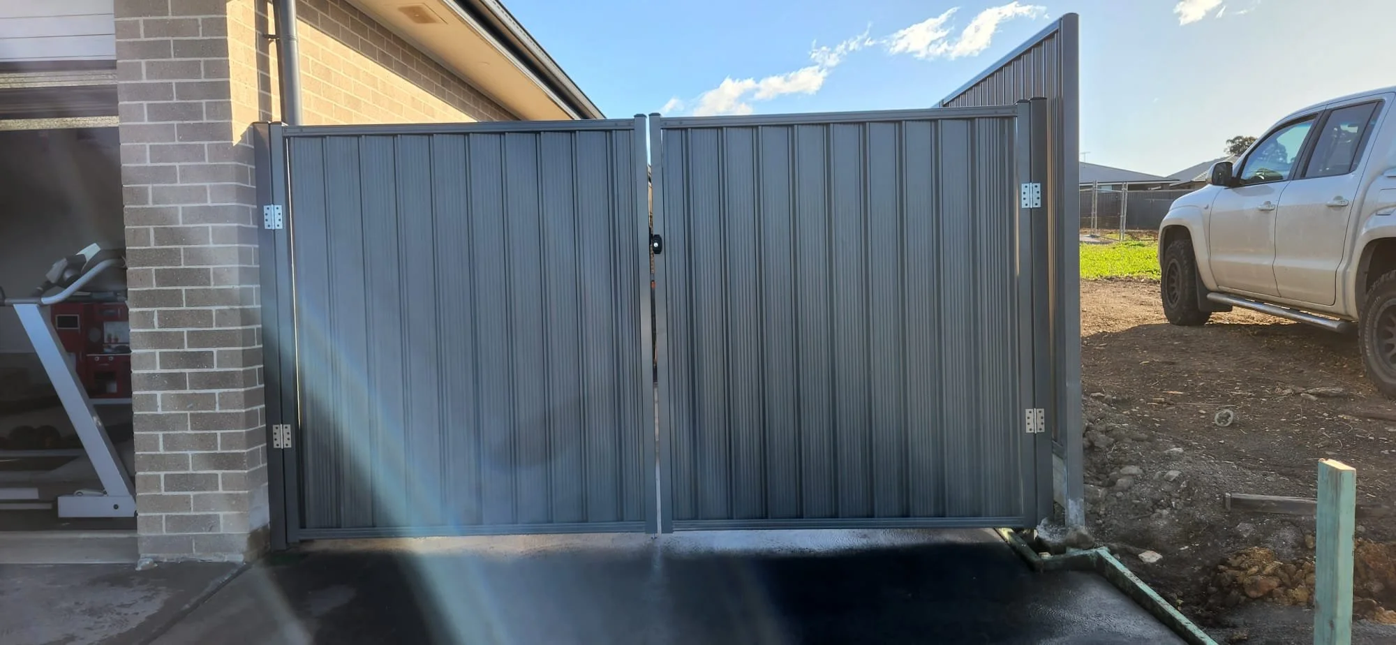 A gray metal gate attached to a brick house with a garage on the left and a car parked on a dirt driveway on the right, under a partly cloudy sky.