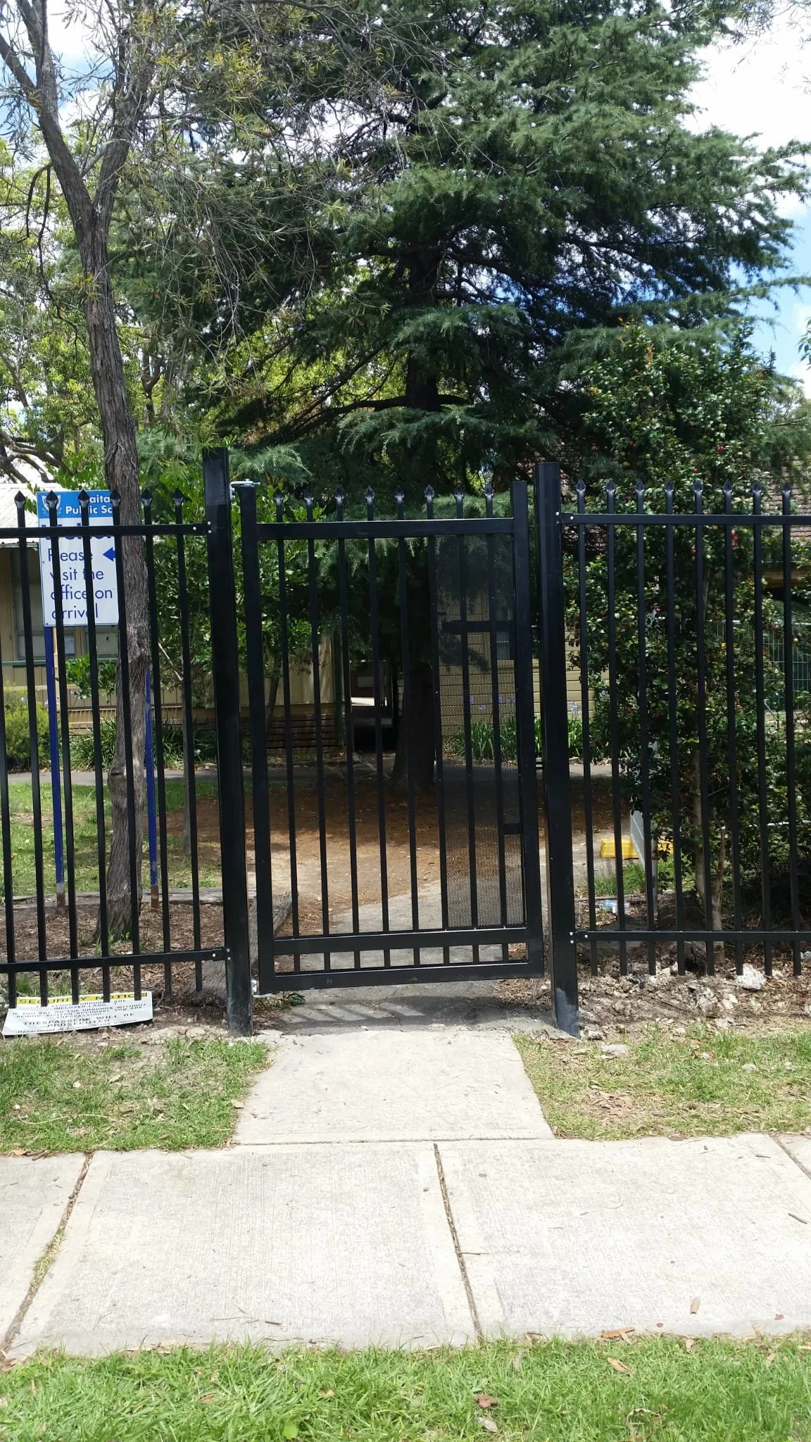 A black metal gate with a screen door at the entrance to a park or school yard, surrounded by green trees and a sidewalk in front.