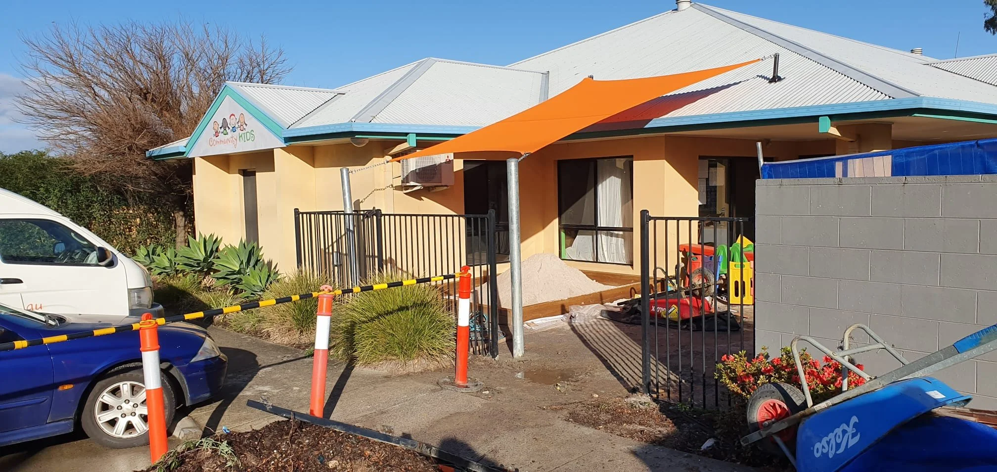 Exterior of a children's community center with construction work, including parked cars, construction barriers, and tools, under a blue sky.