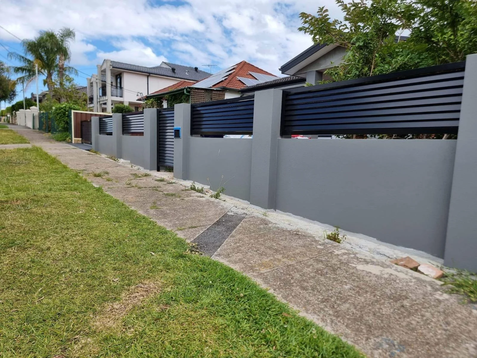 A modern concrete fence with black metal slats runs alongside a sidewalk in a suburban neighborhood, with houses and trees behind it under a cloudy sky.