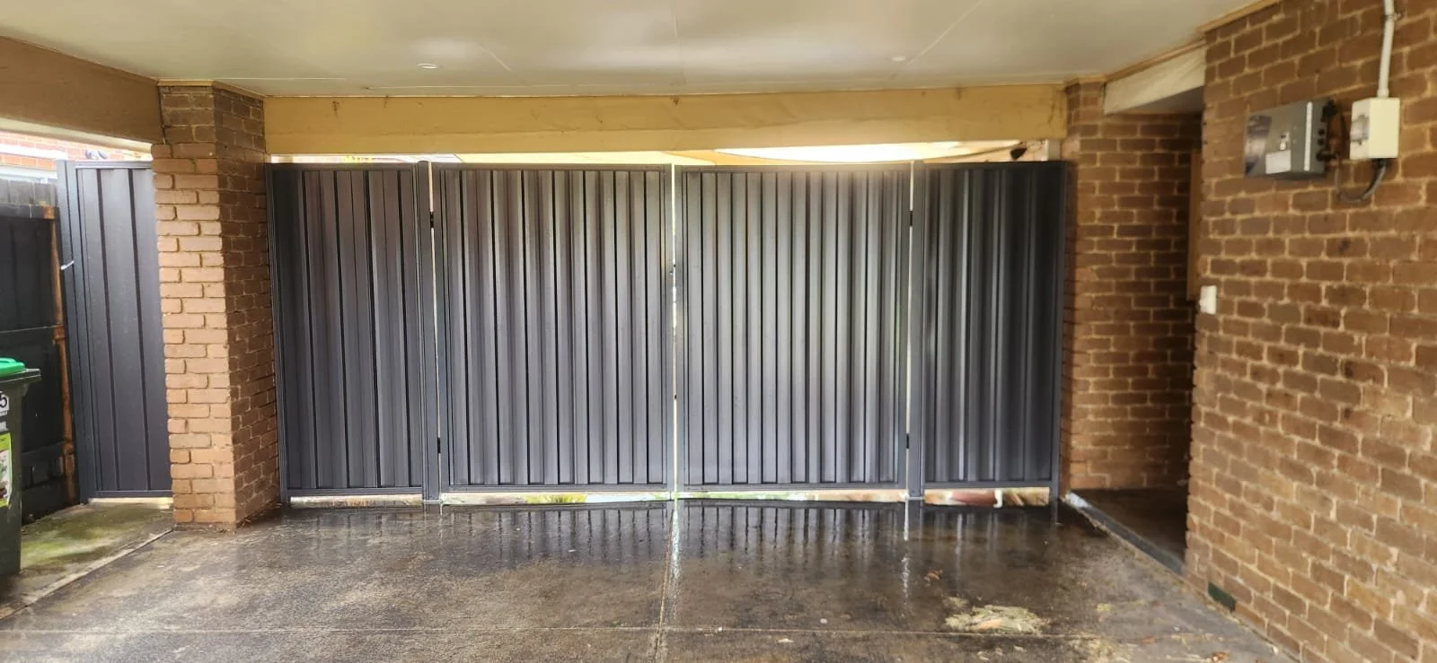 A covered outdoor area with a wet concrete floor, brick walls, and a metal sliding gate. There is also electrical equipment mounted on the right brick wall.