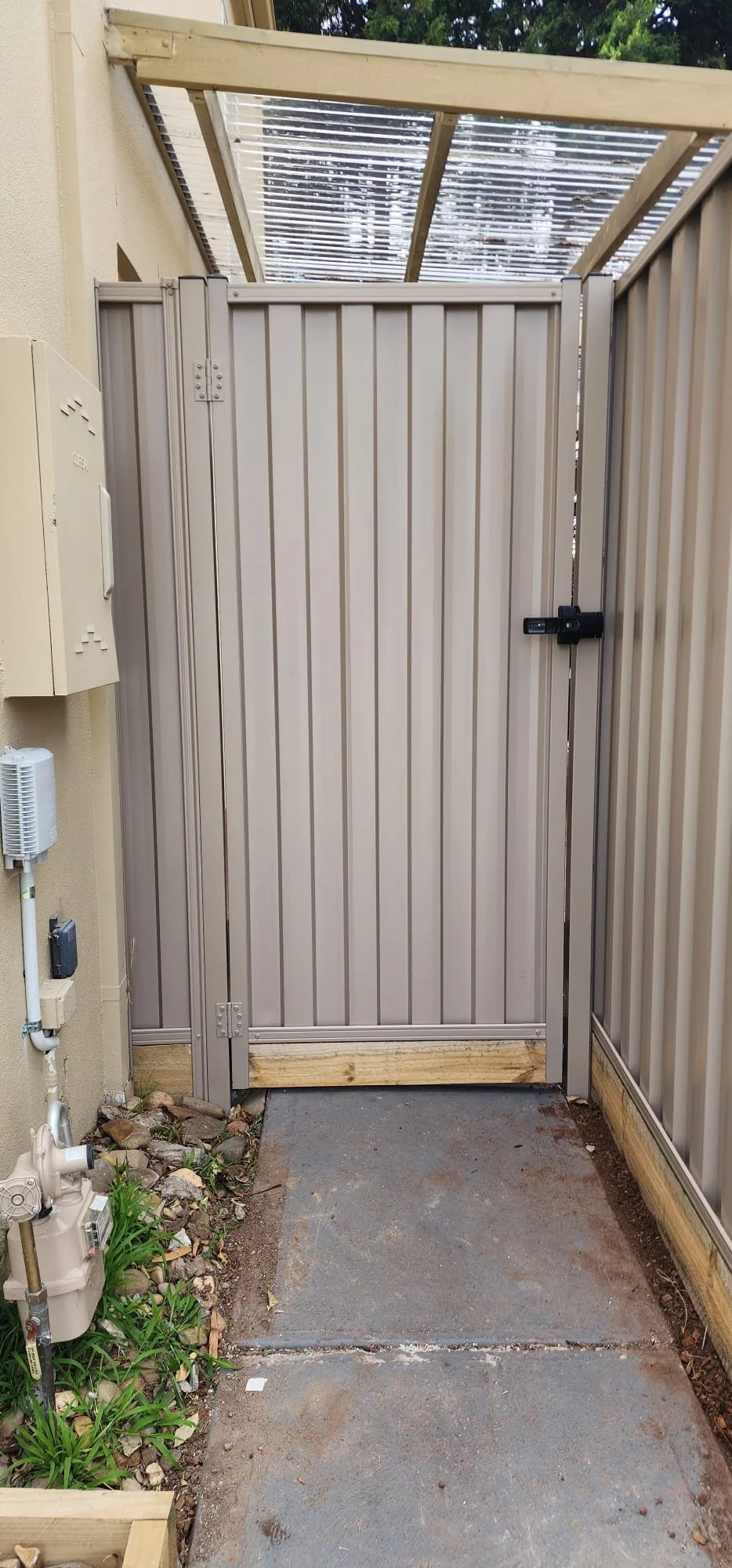 An outdoor enclosed area with a metal gate, wooden frame, and concrete floor, adjacent to a beige building with utility meters and a small garden with rocks and plants.