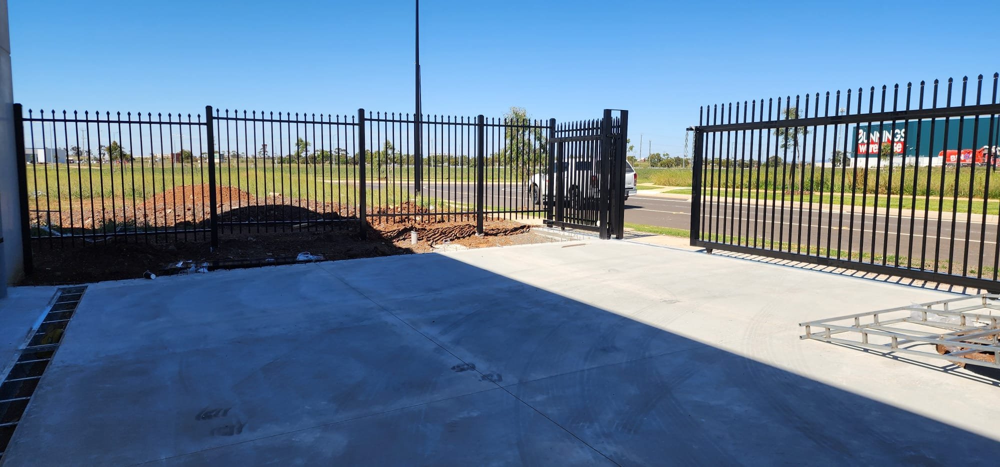 A fenced outdoor area with a concrete patio, black metal fence, and an open gate. Construction material and dirt are visible, with a grassy field, trees, and a road in the background under a clear blue sky.