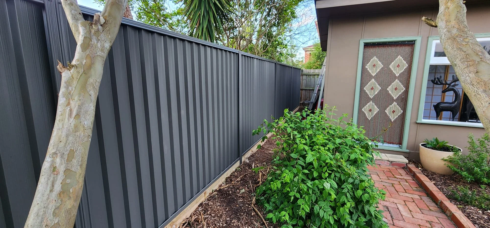 View of a backyard garden with a dark gray metal fence, a brick pathway, green bushes, a beige house with a decorative beaded curtain, a potted plant, and part of a window with a black reindeer figurine inside.
