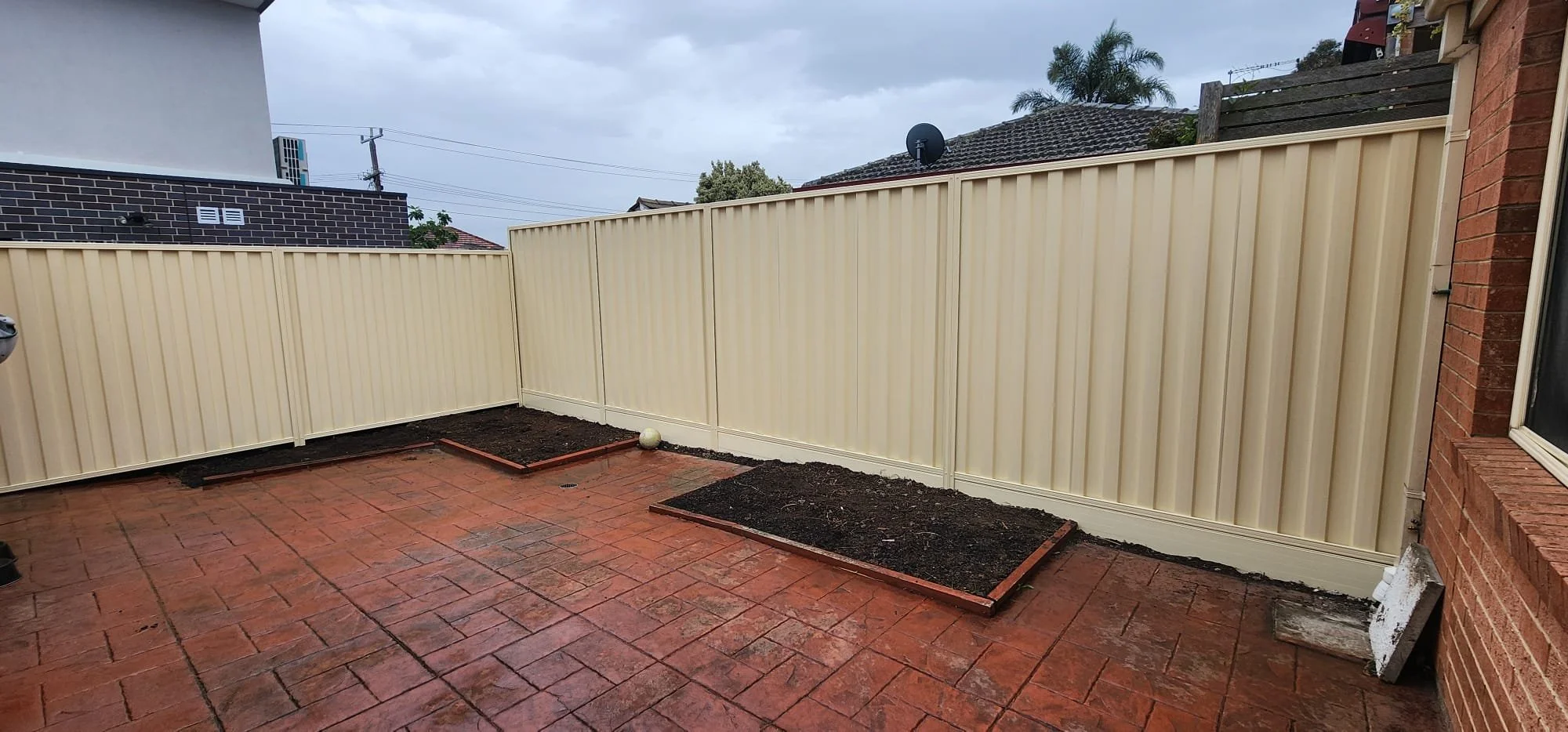 Backyard patio with red brick paving, beige metal fence, and two flower beds with dark soil against the fence.