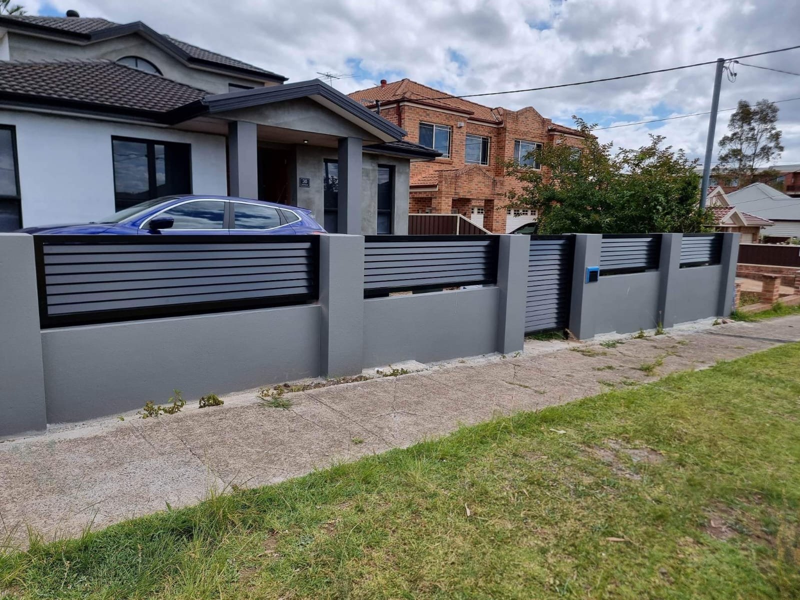 Modern gray fence with horizontal black slats in front of a house with a gray and black exterior. A blue car is parked in the driveway. There are other houses and trees in the background under a partly cloudy sky.