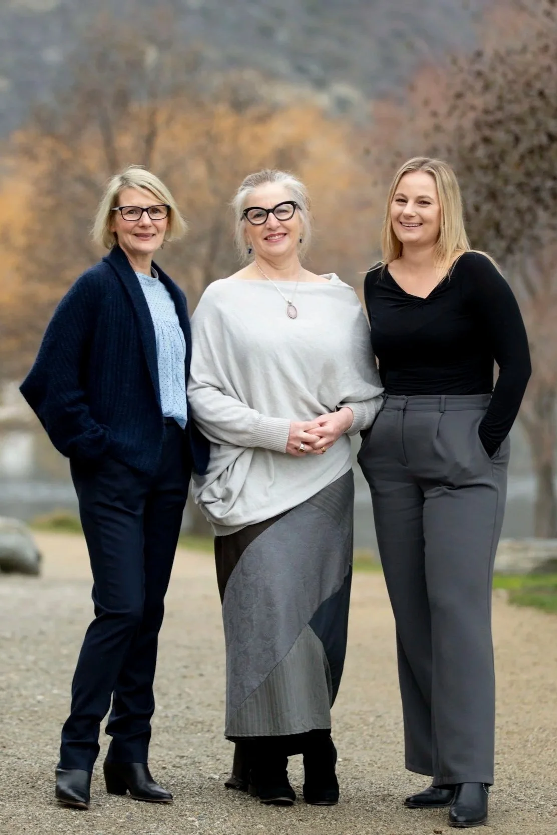 Three women standing outdoors, smiling, with trees in the background.