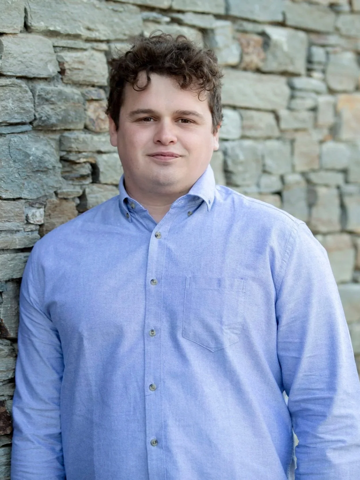 A young man with curly brown hair and a fair complexion wearing a light blue button-up shirt, standing outdoors against a stone wall.
