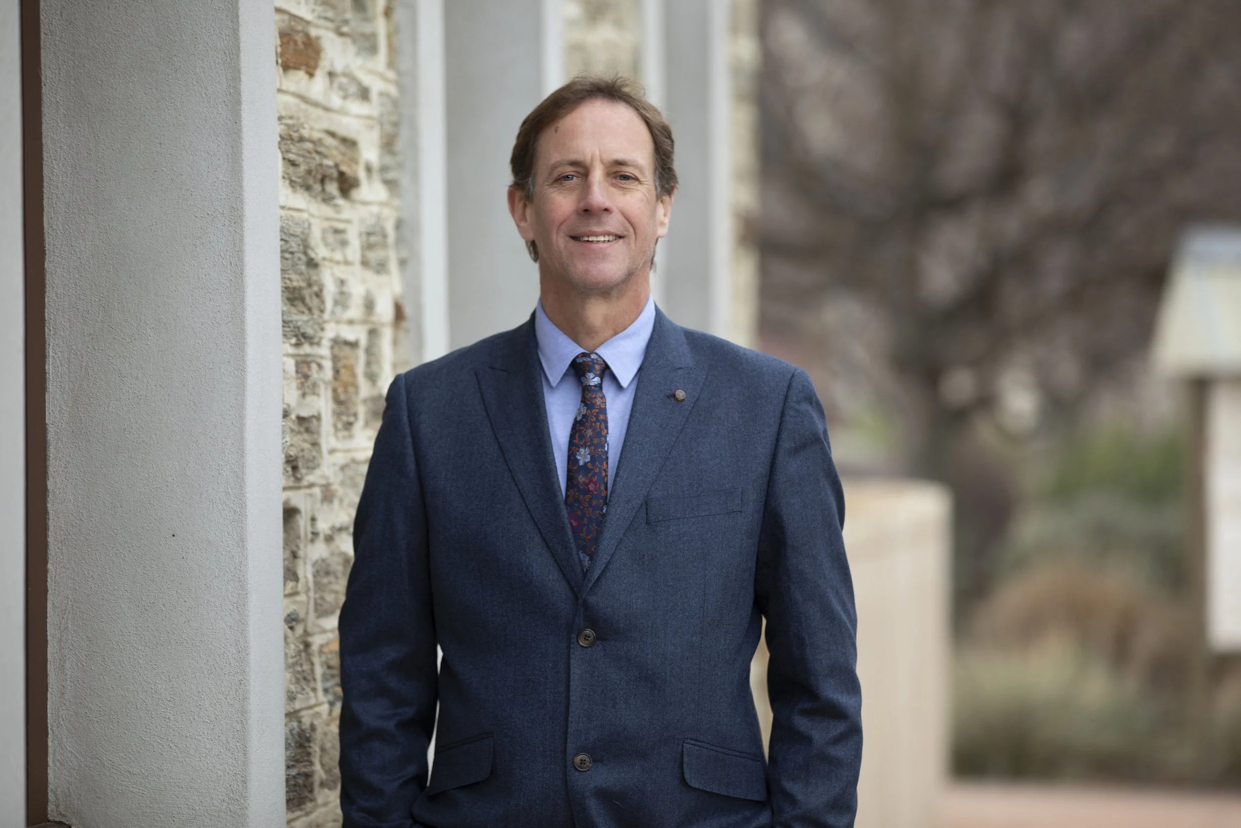 A handsome man in a dark suit with a blue dress shirt standing outdoors against a light-colored wall, looking straight ahead.