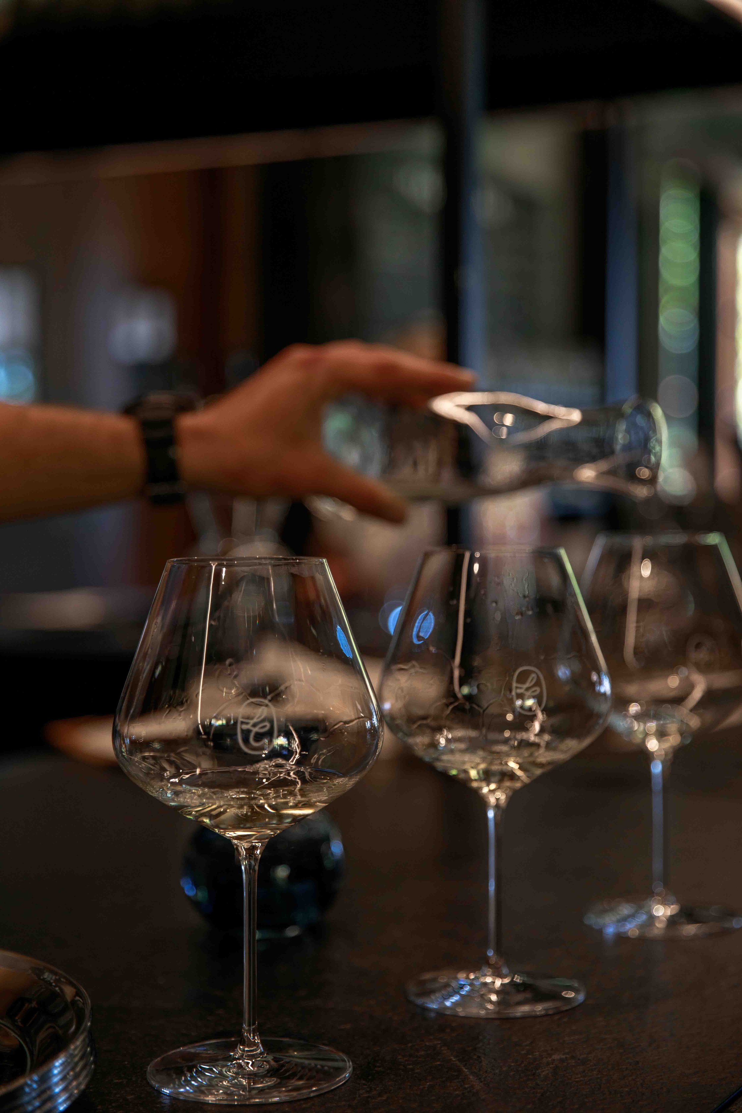 A person reaching to place a glass on a table with several empty wine glasses, in a dimly lit setting with blurred background.