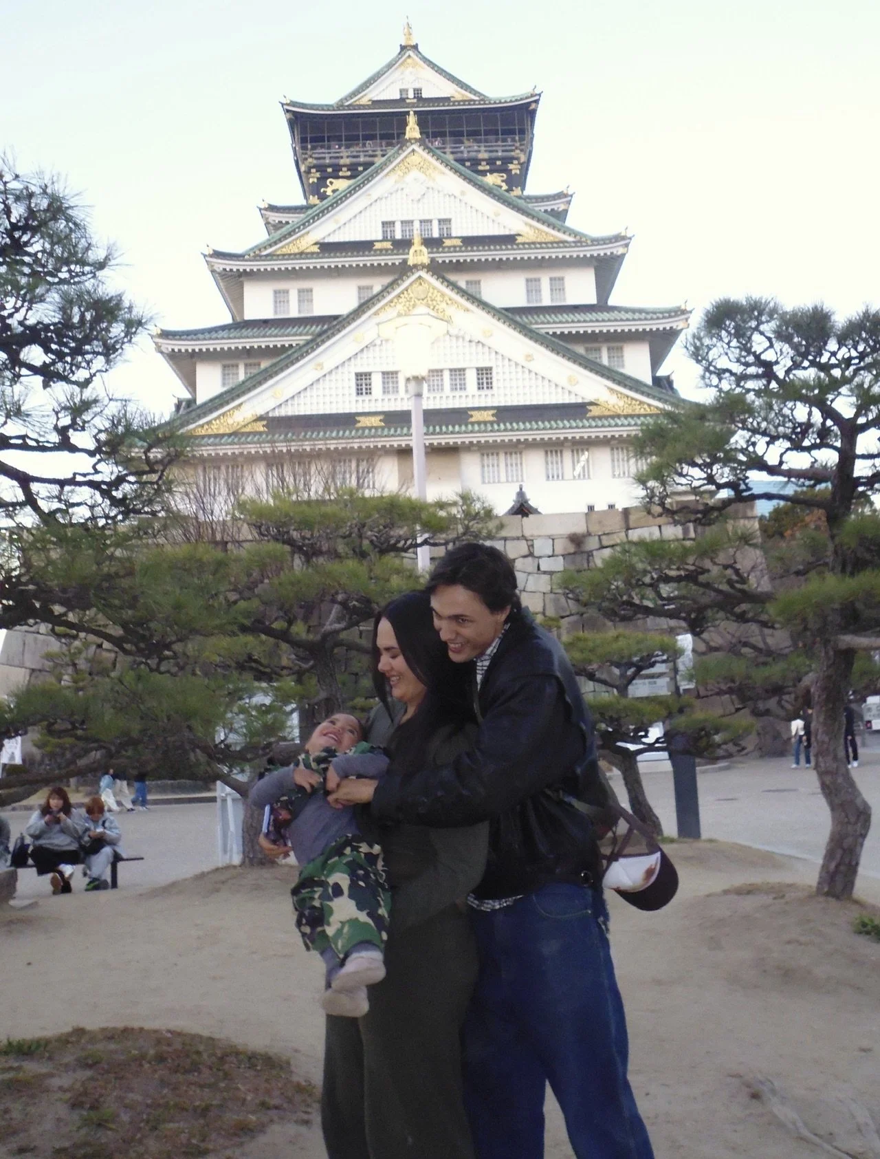 A family of three smiling and playing in front of a traditional Japanese castle with green and white tiers, surrounded by trees and other people.