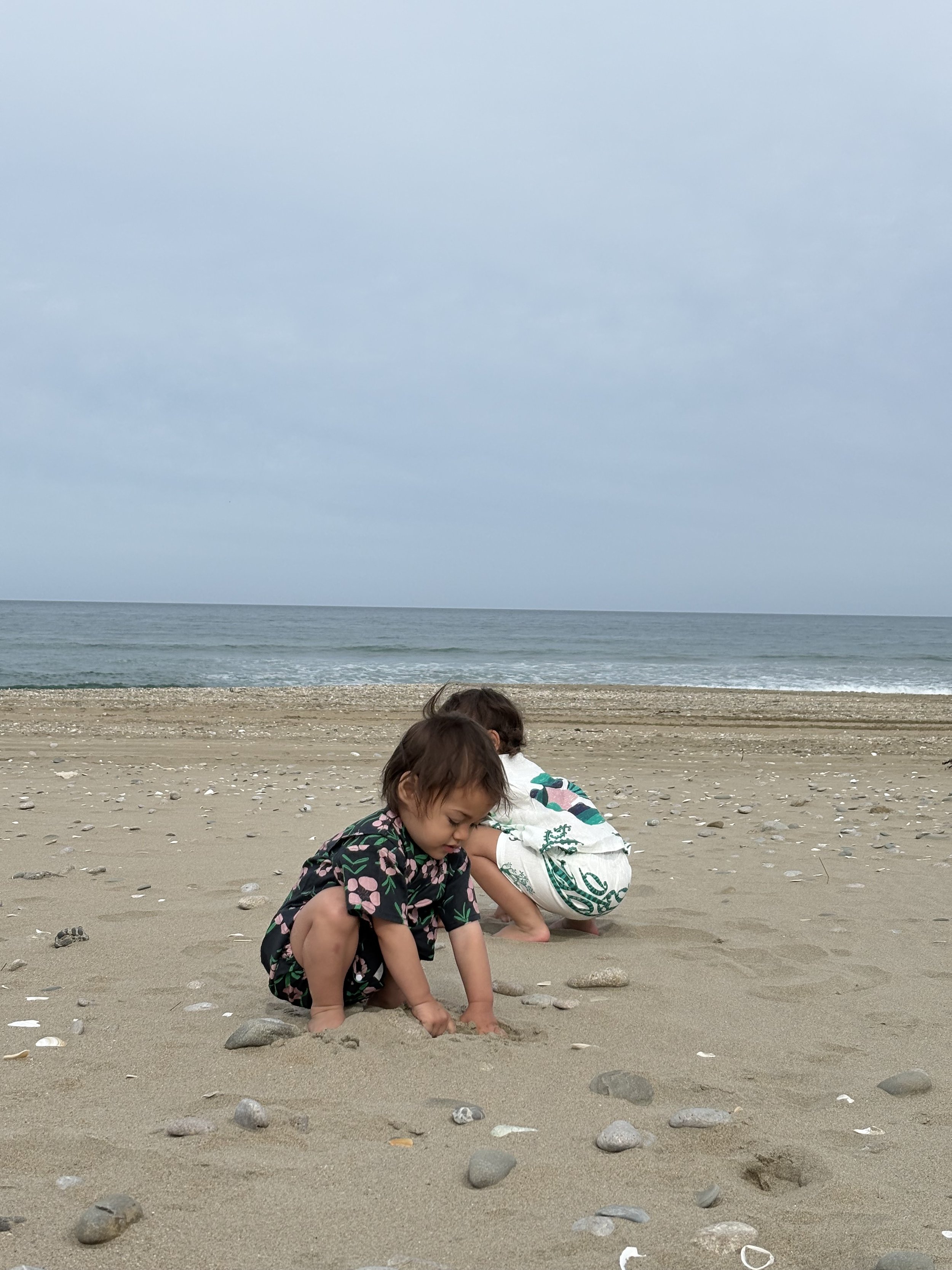 Two young children playing in the sand on a beach, with the ocean and cloudy sky in the background.