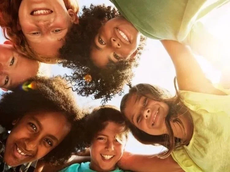 Group of six children looking down at the camera, smiling, with a clear sky in the background.