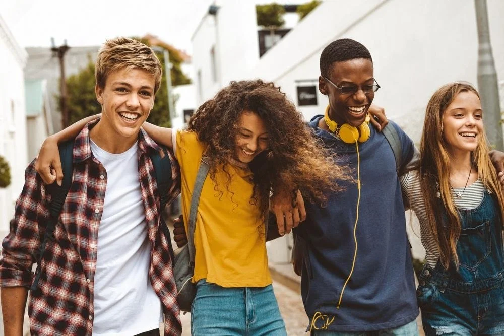 Group of four diverse teenagers walking outdoors with arms around each other, smiling, in a sunny urban neighborhood.