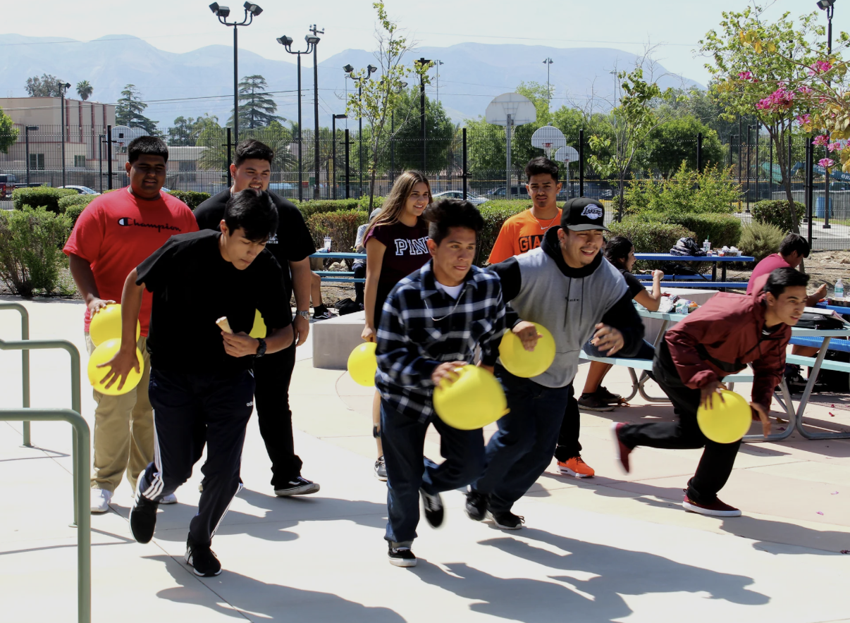 Group of teenagers participating in a balloon race outdoors at a park with trees, benches, and basketball courts, with mountains in the background.