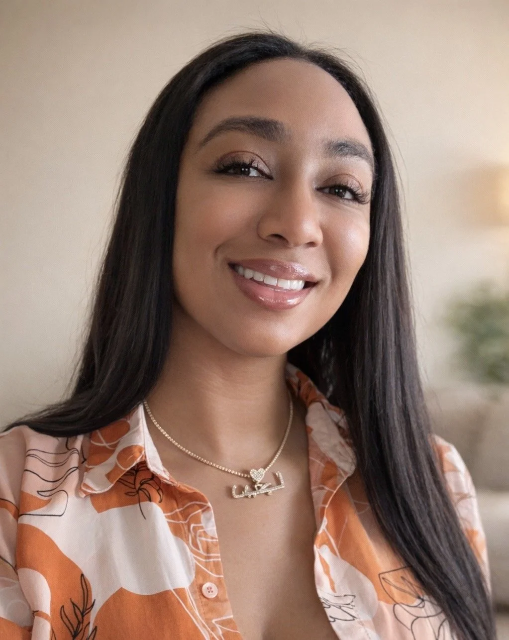 A woman with long dark hair smiling, wearing a patterned orange and white blouse, and a gold necklace with a name and heart charm.