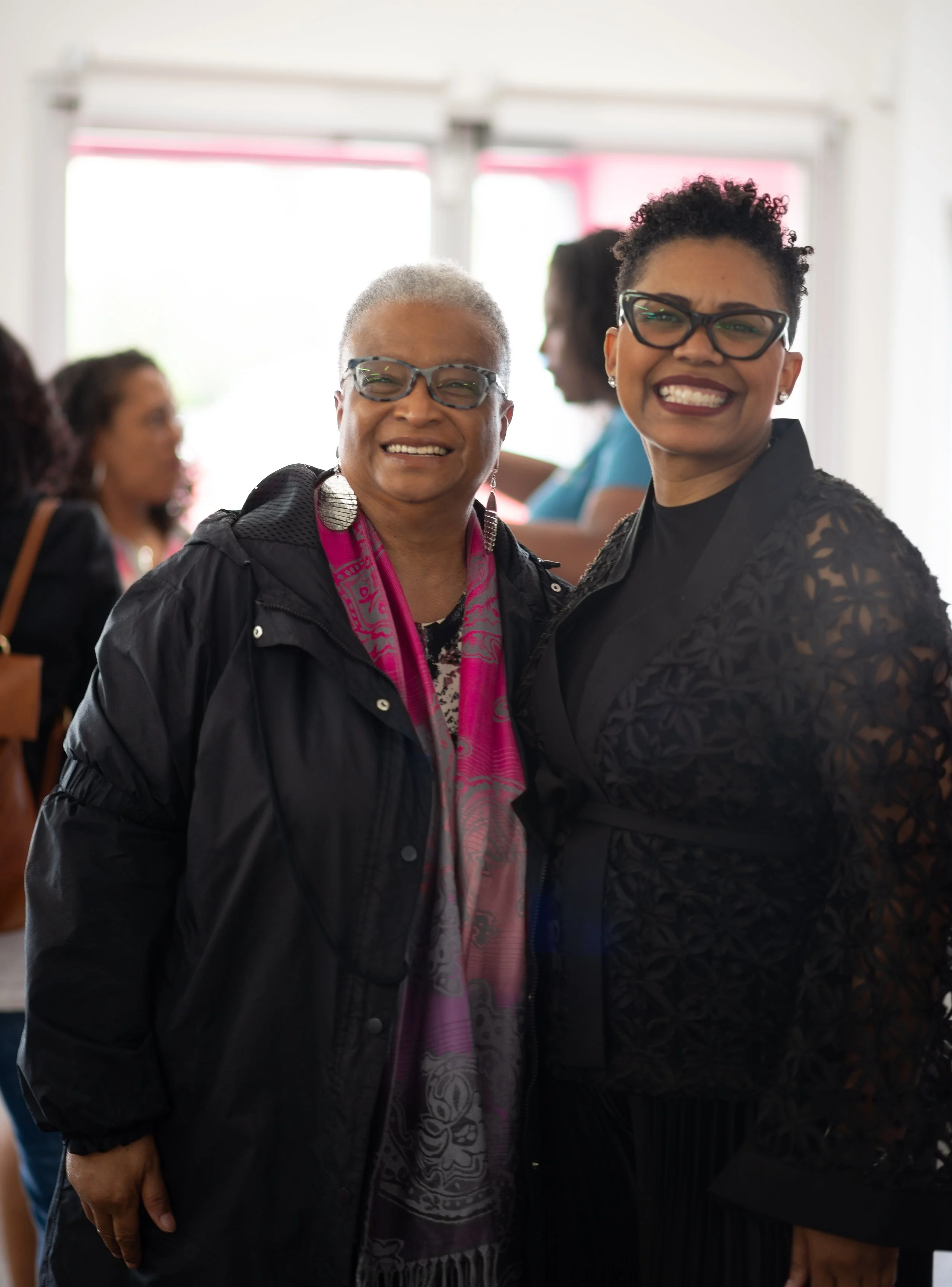 Two smiling women wearing glasses, standing close together indoors with other people in the background.