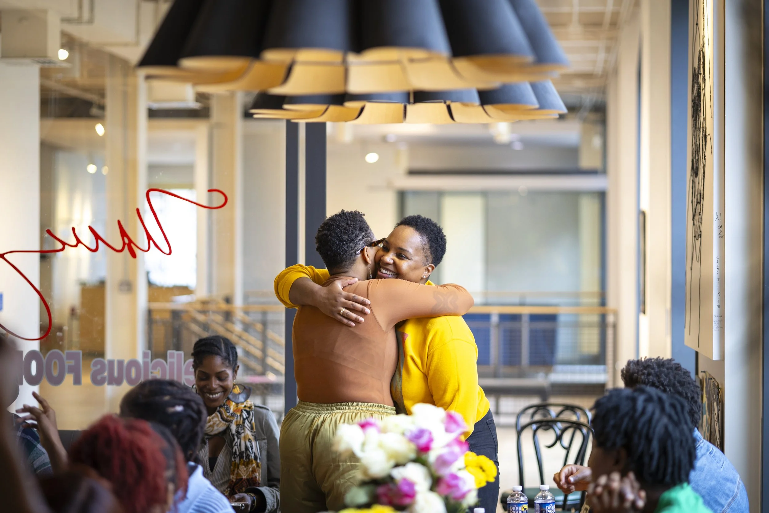 Two women hugging and smiling at each other in a social gathering, with others seated around them and a flower arrangement in the foreground.
