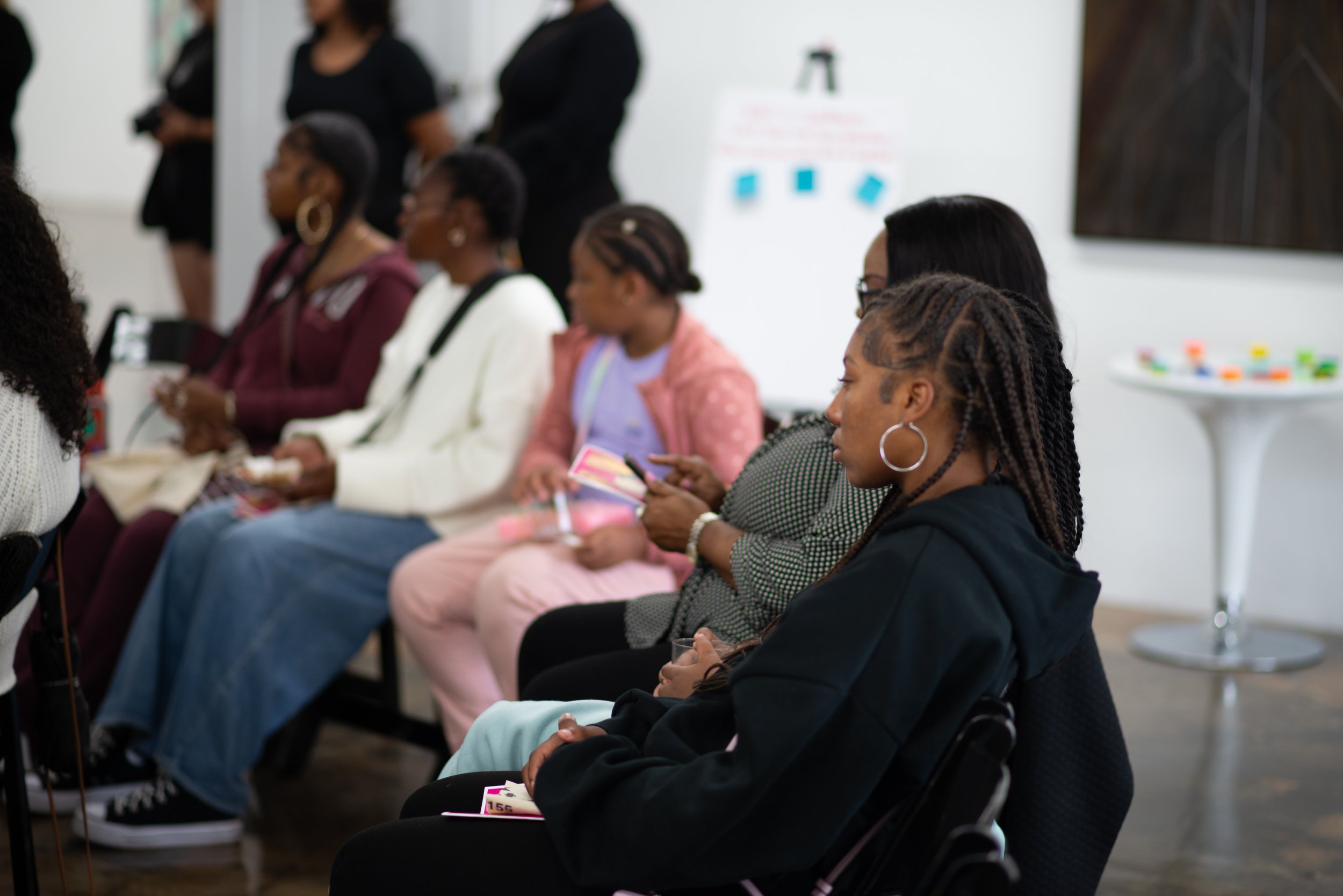 A group of women sitting in a conference room, some are looking at their phones, others are holding notebooks or cups, with a woman standing in the background and a whiteboard on the wall.