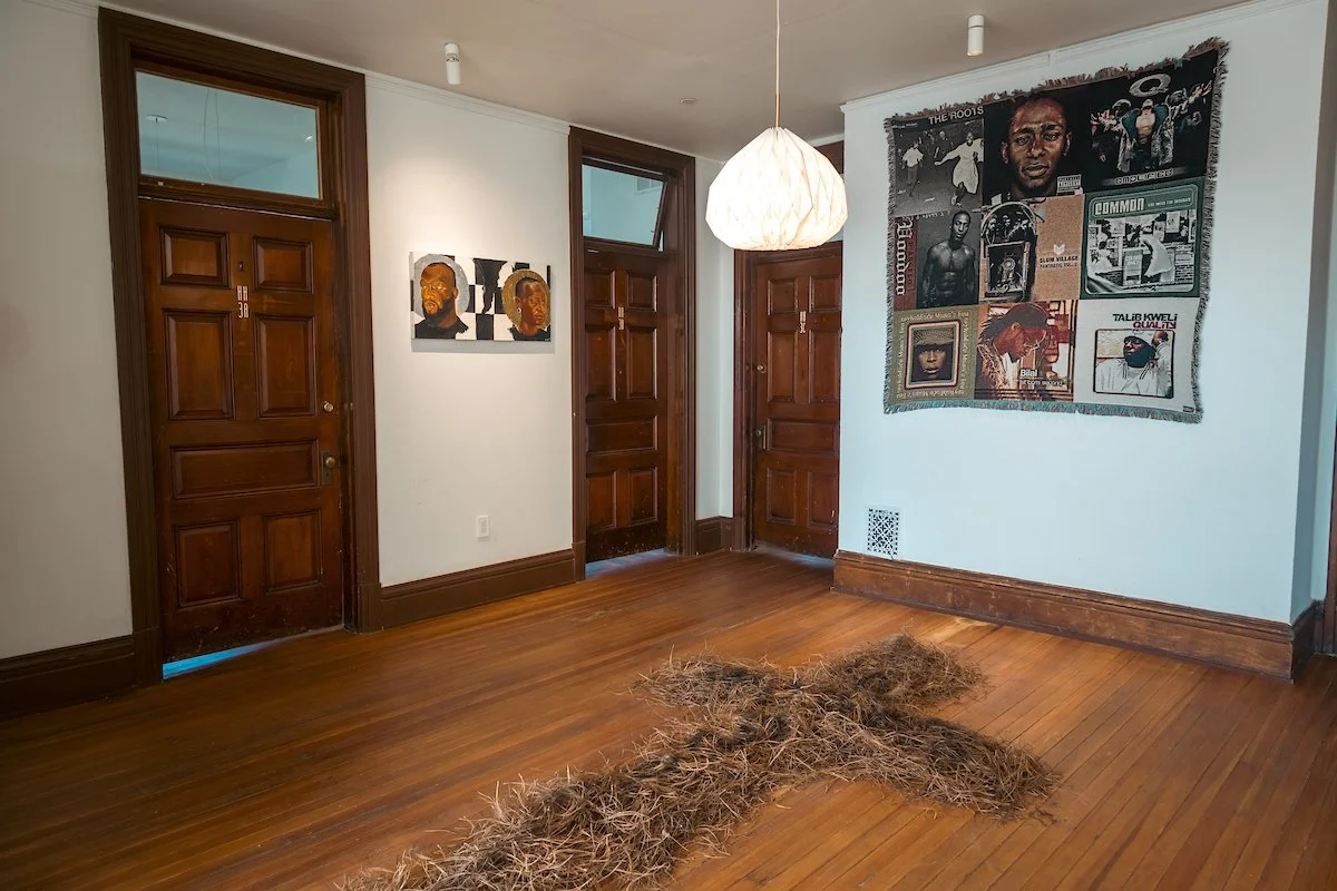 Empty room with hardwood floors, two wooden doors, wall art with portraits, a tapestry, and a pile of dried grass or twigs on the floor.