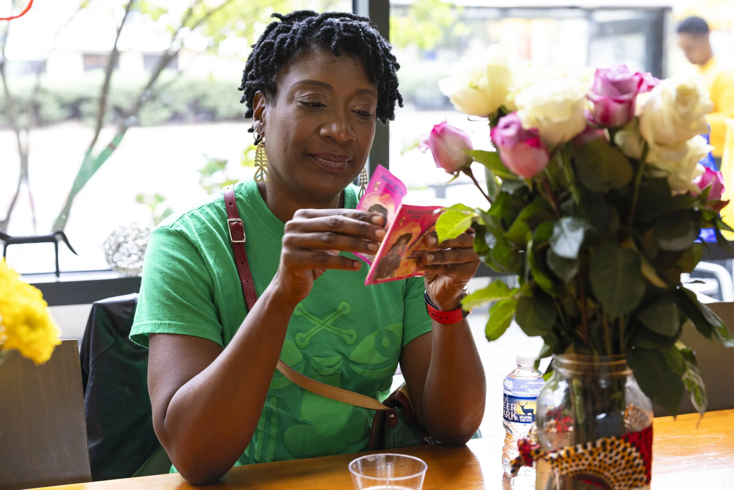 Woman with short black curly hair, wearing a green T-shirt and earrings, sitting at a wooden table, reading pink money, with a vase of pink and white roses and a water bottle on the table in front of her.