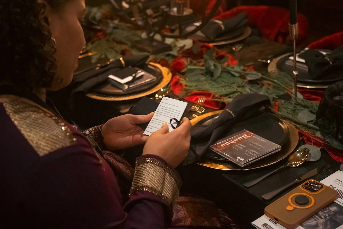 Person sitting at a fancy dinner table reading a menu or program, with place setting, gold spoon, tablet, and phone on the table, decorated with red and green foliage.