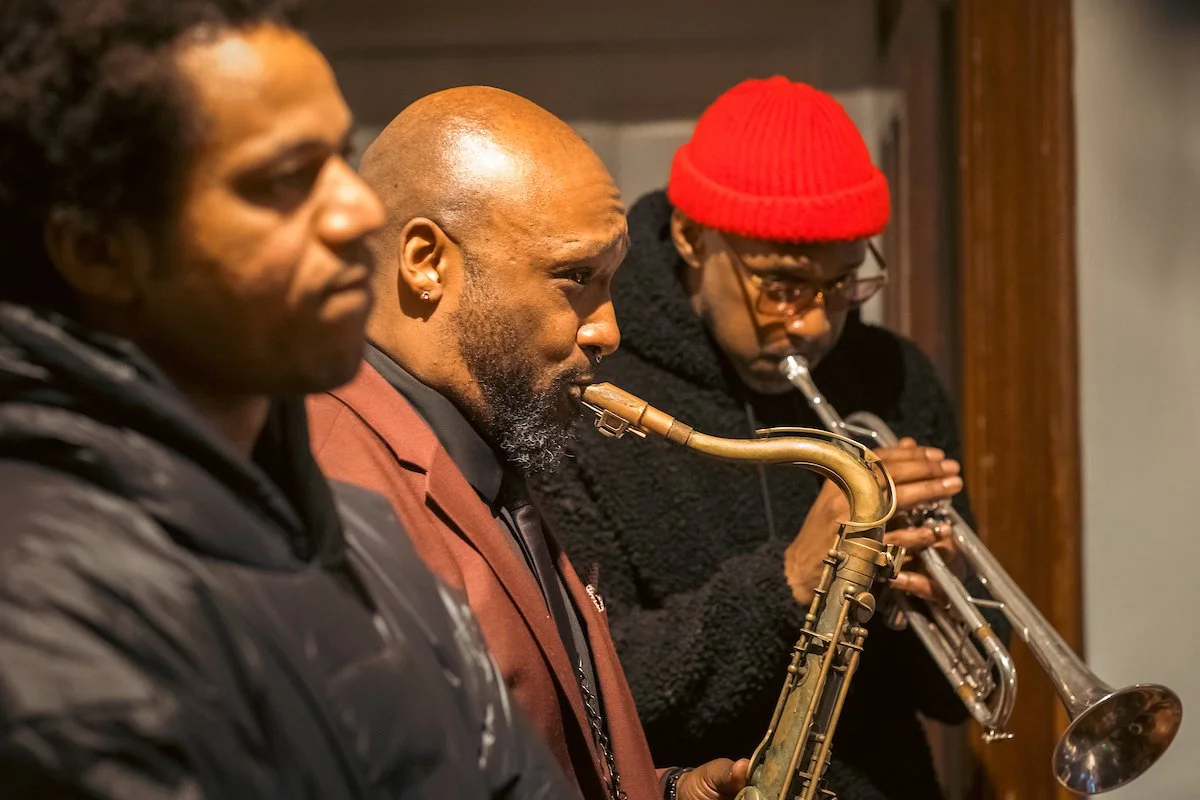 Three musicians, two playing trumpets and one playing a saxophone, in a performing setting.