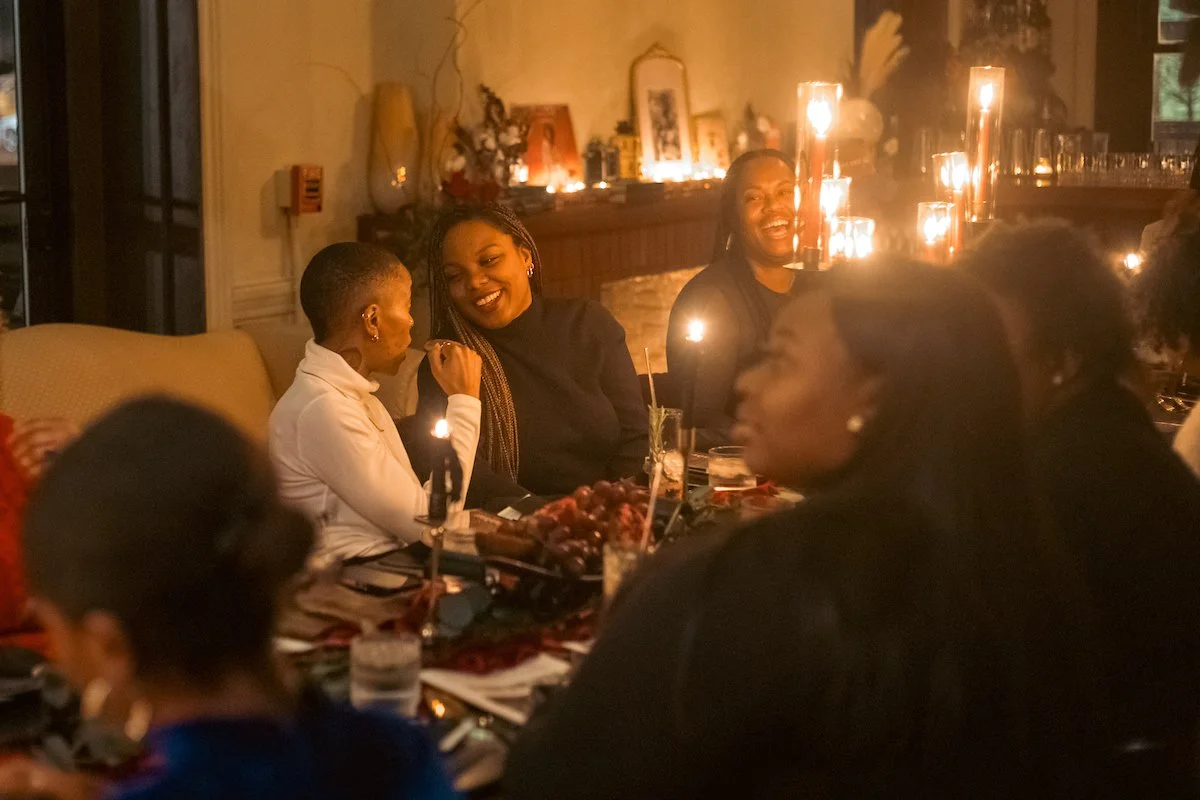Group of women smiling and enjoying a candlelit dinner with food and drinks in a cozy, warmly lit restaurant