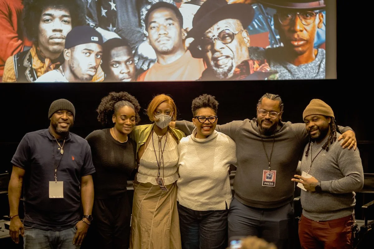 Group of six diverse people standing together with their arms around each other, smiling, in front of a large screen displaying a historical photo of several African American individuals.